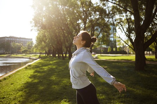 Sideways summer shot of beautiful young woman with hair bun closing eyes, taking deep breath, keeing arms behind, saluting sun while training outdoors in park. Sports, fitness, yoga and meditation