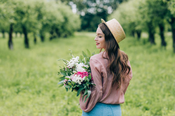 back view of smiling woman in hat with bouquet of flowers in hands standing in park