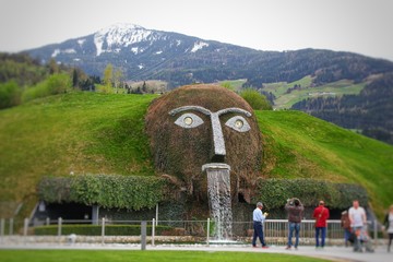 Monster giant head fountain spitting water into a pond at swarovski Kristallwelten in Wattens, Austria with cloudy skies and snowy Alps in background