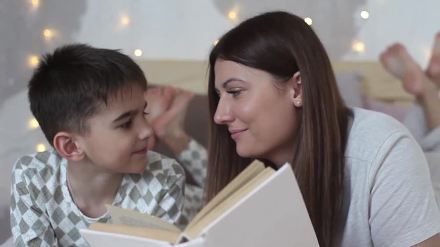 Mom and son are reading a book lying on the bed. Mom kisses her son.