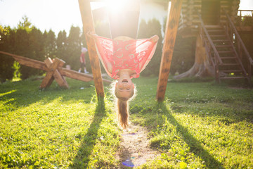 summer children's games and fun outdoors. Happy child girl hanging upside down on wooden Playground...