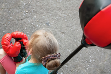 Little girl in red boxing gloves is resting near the punching bag