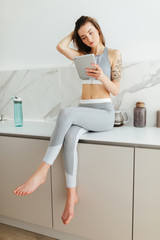 Beautiful girl in sporty top and leggings sitting on kitchen table with laptop in hand and thoughtfully looking aside isolated