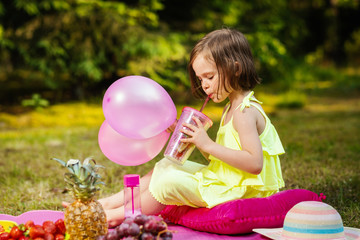 little girl with balloons is drinking lemonade at a picnic in summer forest