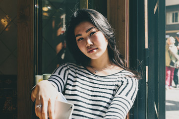 Portrait of a beautiful asian student girl wearing casual outfit smiling at the camera while sitting with a mobile phone in a modern coffee shop. Young blogger having rest in a cafe on a sunny day.