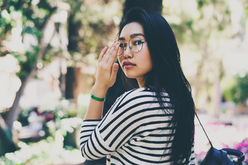 Portrait of a beautiful asian woman with long dark hair wearing glasses with a leather backpack turning back and looking at the camera while walking the city park on a sunny summer day.