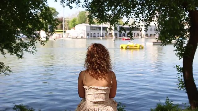 Curly woman sitting and relaxing by lakeside, shot from behind on a summer day