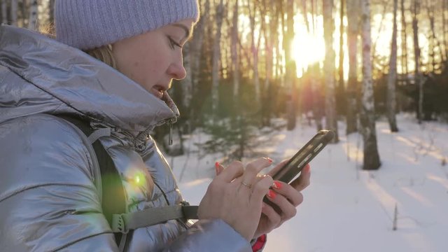 Woman is walking in the woods. Traveler is photographed on the phone in forest. Girl does selfie and communicates with her smartphone. Evening time with beautiful juicy sunset. Explorer is dressed in