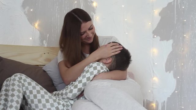 A beautiful mother is playing with her son sitting on the bed having a good mood.