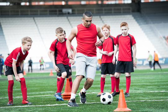 Full length portrait of junior football team practicing in stadium, focus on muscular coach leading ball in foreground, copy space
