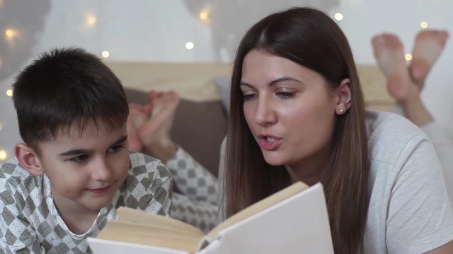A beautiful girl with long hair reads a book to a small boy lying on a bed and kisses him having a good mood.HD