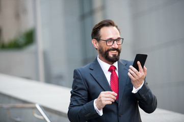Businessman in glasses using smart phone on office walkway with city building background. Concept of business people using technology. man in suit is searching information on web via mobile phone.