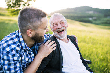 An adult hipster son with senior father in wheelchair on a walk in nature at sunset, laughing.