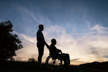 A silhouette portrait of adult son with senior father in wheelchair in nature at sunset.