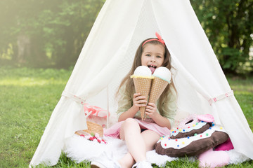 Little girl playing in a tent, children's house wigwam in park. Happy mothers day. Child with big donuts and ice cream and straw hat. Summer, happy childhood concept. Cute emotions girl. © MartaKlos
