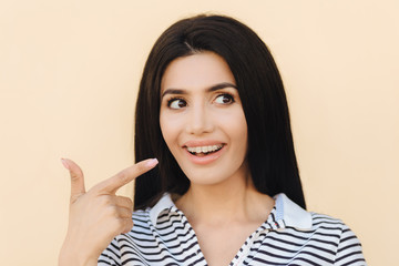 Pretty young brunette female with healthy skin, indicates at teeth with braces, wears striped t shirt, looks happily aside, shows her beauty, has make up, isolated over light studio background