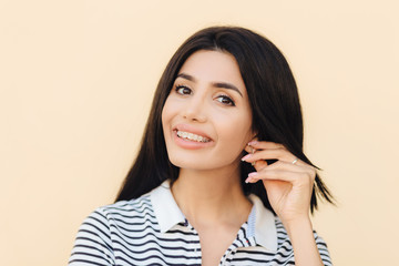 Fototapeta premium Headshot of cute lovely woman has broad smile, dark hair, keeps hand on ear, shows white teeth with braces, looks directly into camera, has happy expression, isolated over studio background.