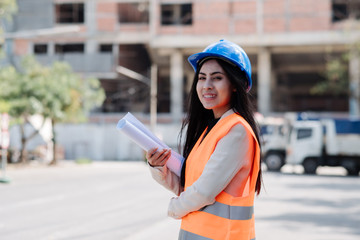 Asian woman engineer with the blue safety helmet meeting at the construction site with blueprints and laptop.