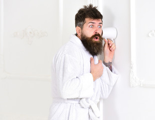 Man with beard and mustache eavesdrops using cup near wall. Hipster in bathrobe on surprised face...
