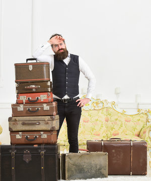 Macho elegant on tired face, exhausted at end of packing, leans on pile of vintage suitcases. Baggage and relocation concept. Man with beard and mustache packed luggage, white interior background.
