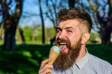 Chilling concept. Man with long beard licks ice cream, close up. Bearded man with ice cream cone. Man with beard and mustache on happy face eats ice cream, nature background, defocused.