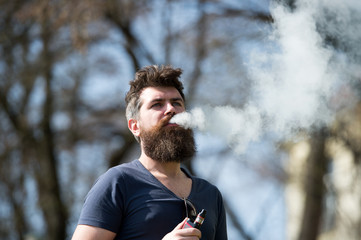 Electronic cigarette concept. Man with long beard and clouds of smoke looks relaxed. Bearded man smokes vape on sunny day. Man with beard and mustache on calm face, branches on background, defocused