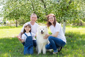 Happy family with a white dog in a summer park. The husband, wife and little daughter in white clothes and jeans, along with a white dog, rest in the garden.