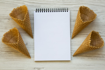 Waffle sweet cones with notepad on a white wooden background, top view. From above. Copy space.
