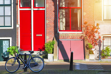 Fototapeta na wymiar Picturesque in Netherlands. Bicycle near old house with red door and window at the canal with sunlight in Amsterdam, Nord Holland, Netherlands.