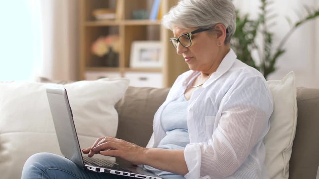 technology, old age and people concept - happy senior woman in glasses typing on laptop at home
