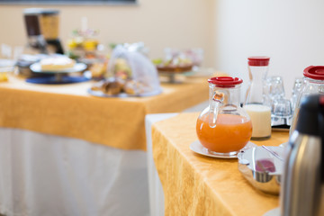 Table with buffet breakfast. Glass bottle with milk and glass pitcher with orange juice. Blurred table with food in the background