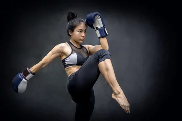 Papier Peint photo Lavable Arts martiaux Jeune boxeuse asiatique avec des gants de boxe bleus donnant des coups de pied dans la salle d& 39 exercice, arts martiaux sur fond noir