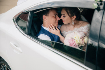 The bride and groom are sitting in the back seat of the car, the girl is holding a bouquet of flowers in her hands. Lovers almost touch each other with their noses, hug and kiss.