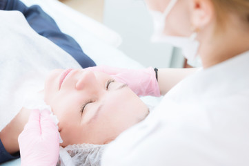 Fototapeta premium A close-up of the cleaning procedure in the office of cosmetology. The hands of the cosmetologist in pink gloves are removed from the face of a young girl with a sponge cleansing the mask