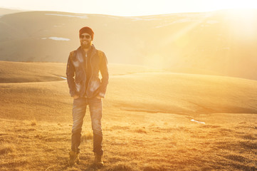 Fashion portrait of a bearded hipster young man wearing sunglasses, a backpack and hat on a background with copyspase in the mountains at sunset .. A confident man with a beard.