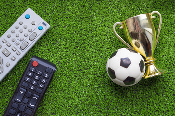 Naklejka premium TV remote control and soccer ball with gold cup on the green grass. program championship football concept.