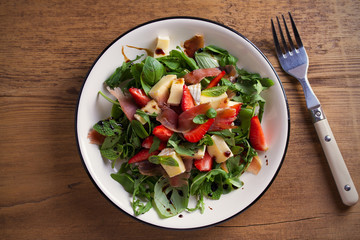 Strawberry, basil, mint, arugula salad with brie cheese and jamon on white plate on wooden table. horizontal, overhead