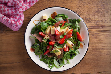 Strawberry, basil, mint, arugula salad with brie cheese and jamon on white plate on wooden table. horizontal, overhead