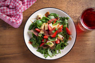 Strawberry, basil, mint, arugula salad with brie cheese and jamon on white plate on wooden table. horizontal, overhead