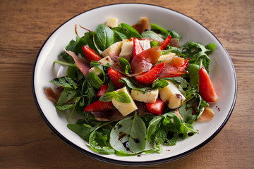 Strawberry, basil, mint, arugula salad with brie cheese and jamon on white plate on wooden table. horizontal