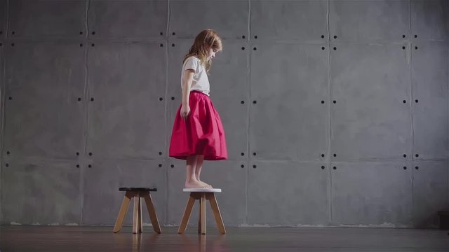 Side view of an adorable little girl wearing a pink skirt walking on small wooden chairs in a gray wall room. Locked down slow motion medium shot