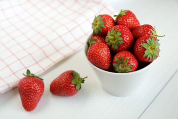 Red ripe strawberries in a cup and checkered dish towel on white wooden table top view.