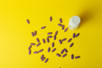 a white box for tablets stands on a yellow background, beside lies a bunch of oblong, purple pills.