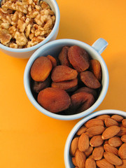 Diagonal closeup of healthy snacks, dried apricots, walnut halves and almonds, in blue bowls on orange background.