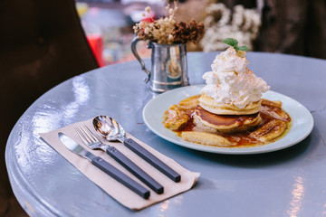Pancakes with caramel-banana syrup topping with whip cream and mint leaves. With spoon, fork and knife.