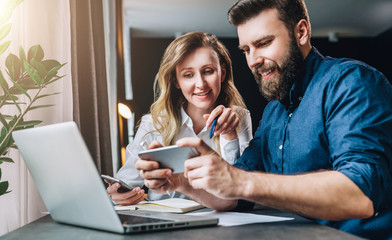 Business meeting. Teamwork. Smiling businesswoman and businessman sitting at table in front of laptop. Man showing woman information on smartphone screen. Online marketing, e-learning, distance work.