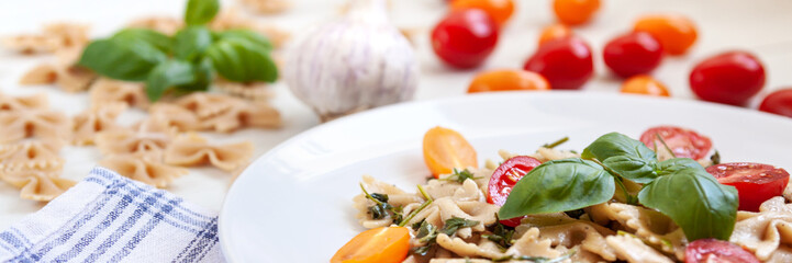 Panoramic view on white plate with farfalle aglio olio, cherry tomatoes, fresh basil and garlic