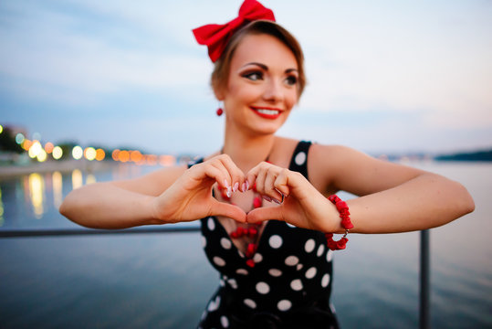 A beautiful young woman dressed stand on the pier in the evening, shows with his own hands the heart