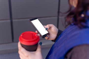 close-up of female hands holding coffee and smartphone with blank screen for your logo or text message, girl walking around town at night and drinking cappuccino