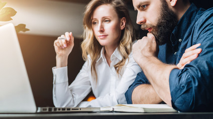 Business meeting. Teamwork. Businesswoman and businessman sitting at table in front of laptop and working. Couple of millennials pondering business project. Online education, e-learning, e-commerce.